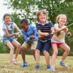 A group of children pulling a rope in a lively park, surrounded by grass and trees.